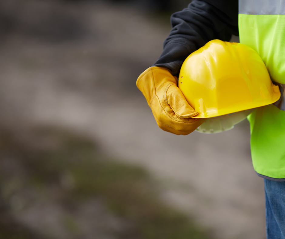Person stands off camera. Only their mid-section is visible. They are wearing yellow gloves, a black sleeve top, jeans, high-vis vest and they are holding a yellow hard-hat. 