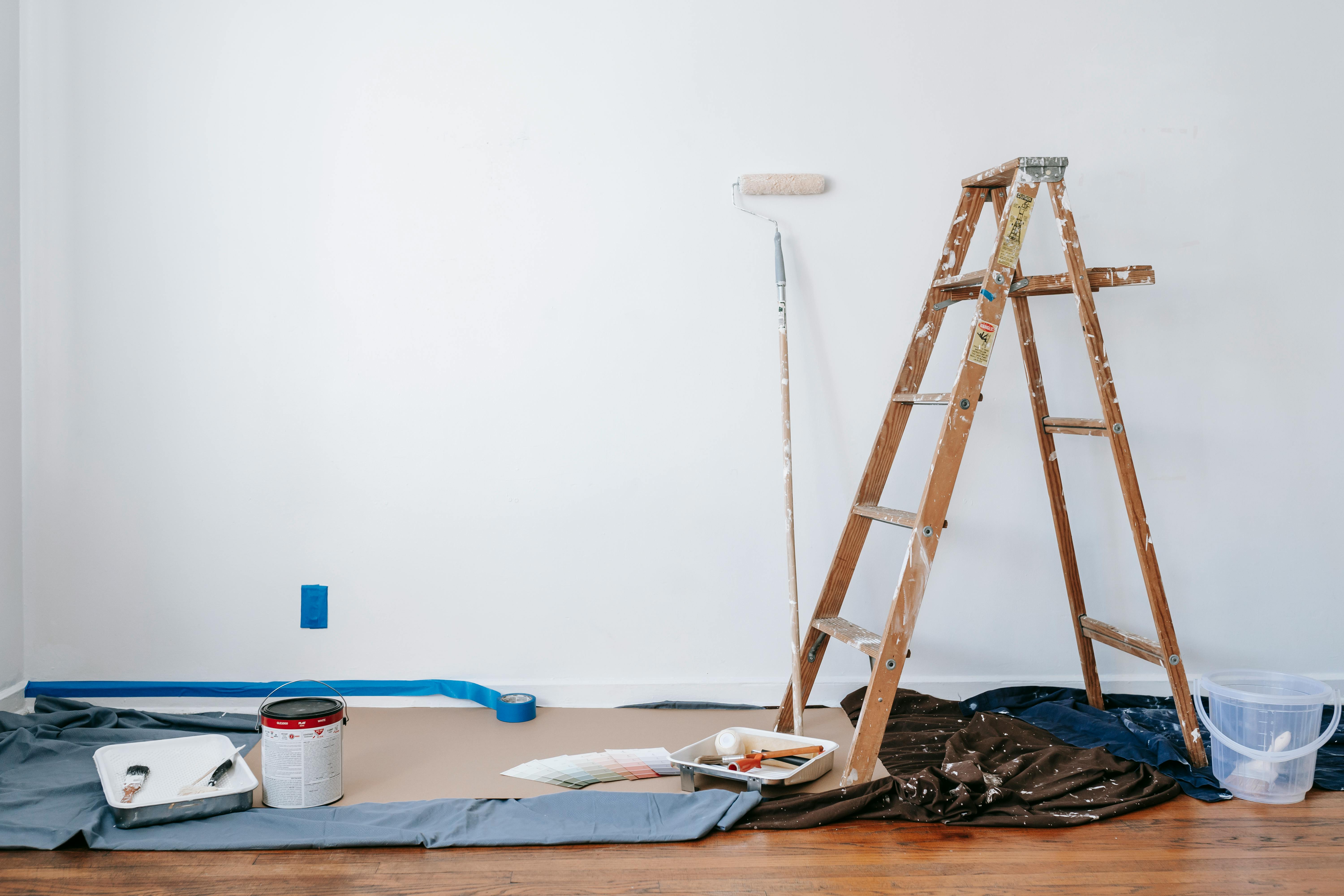 A room with a white wall, in front of it is a ladder, tall paint roller brush, tin of paint and sheets on the floor. 
