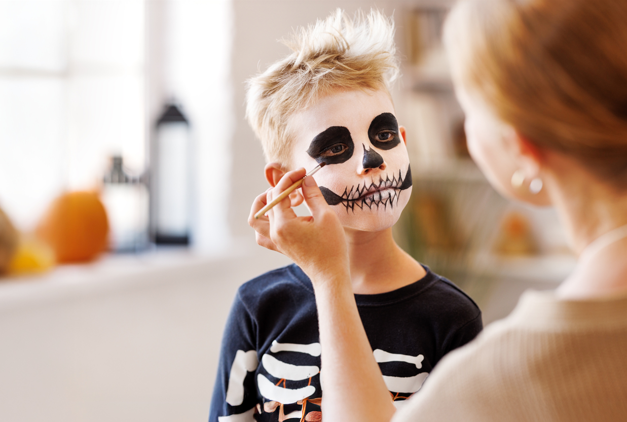 A person is applying face paint to a child. The child has a painted skull face design on the cheek and forehead and is wearing a black skeleton T-shirt.